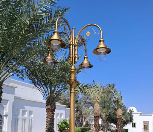 Богато украшенный уличный фонарь в золотом абажуре с четырьмя светильниками 04/Aries/A, на фоне пальм и белых зданий в солнечный день